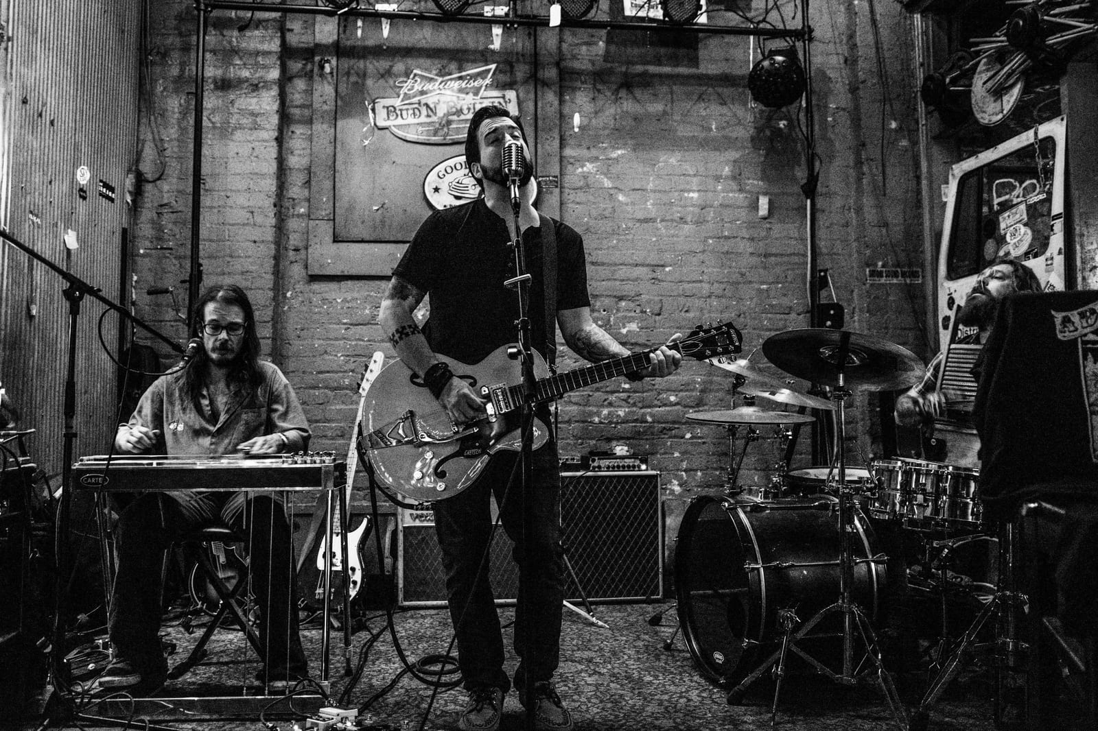 Aden Paul and band on stage — guitar, pedal steel, and drums against a brick wall