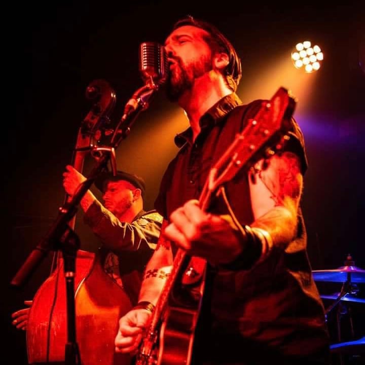 Aden Paul singing into a vintage microphone under amber stage lights, upright bassist behind him